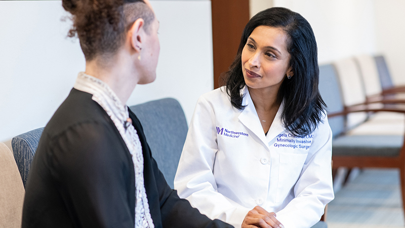 Angela Chaudhari, MD, wearing a white lab coat with NM logo, her name, and Minimally Invasive Gynecologic Surgery printed on it, in a waiting room speaking with a patient whose back is to the camera.