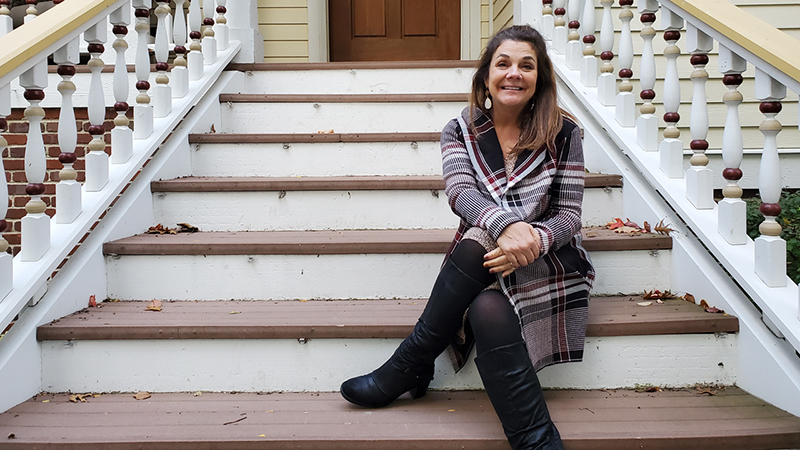 Wendy Hennessey is pictured sitting on the steps of a pale yellow Victorian style house. She has long brown hair and wears a plaid coat and knee-high black boots.