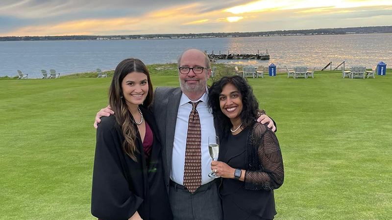 Mark Dybas wears glasses, shirt, a tie and blazer. He is pictured center with his wife and daughter on either side of him. They are all on a green lawn with a lake and sunset behind them.