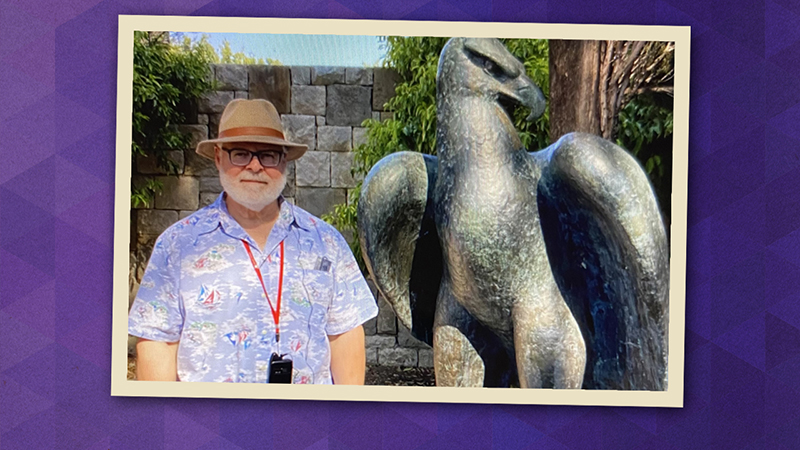 Michael Bemi in a colorful shirt and tan fedora hat, next to a bronze sculpture of an eagle.